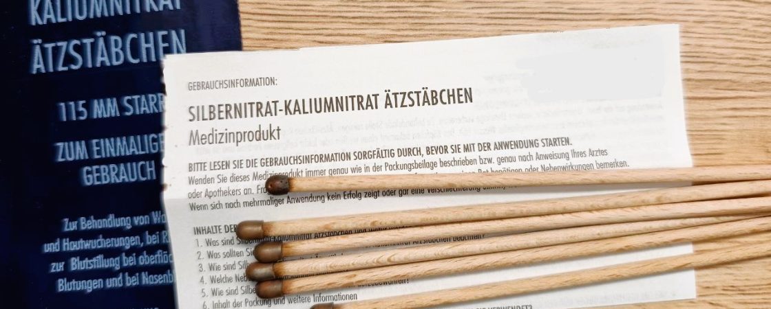 Mehrere Silbernitrat-Kaliumnitrat Ätzstäbchen aus Holz mit dunkler Spitze liegen auf einer Holzoberfläche. Daneben befinden sich die Verpackung und eine Gebrauchsinformation, die Hinweise zur Anwendung und Sicherheit des Medizinprodukts gibt.