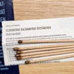 Mehrere Silbernitrat-Kaliumnitrat Ätzstäbchen aus Holz mit dunkler Spitze liegen auf einer Holzoberfläche. Daneben befinden sich die Verpackung und eine Gebrauchsinformation, die Hinweise zur Anwendung und Sicherheit des Medizinprodukts gibt.