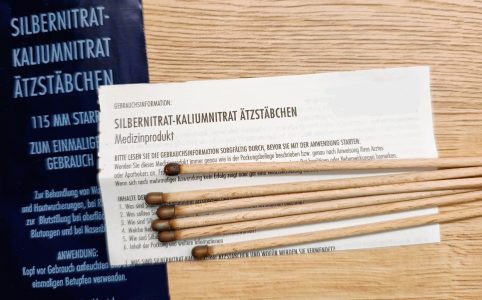Mehrere Silbernitrat-Kaliumnitrat Ätzstäbchen aus Holz mit dunkler Spitze liegen auf einer Holzoberfläche. Daneben befinden sich die Verpackung und eine Gebrauchsinformation, die Hinweise zur Anwendung und Sicherheit des Medizinprodukts gibt.