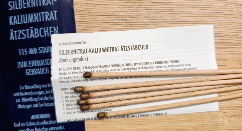 Mehrere Silbernitrat-Kaliumnitrat Ätzstäbchen aus Holz mit dunkler Spitze liegen auf einer Holzoberfläche. Daneben befinden sich die Verpackung und eine Gebrauchsinformation, die Hinweise zur Anwendung und Sicherheit des Medizinprodukts gibt.