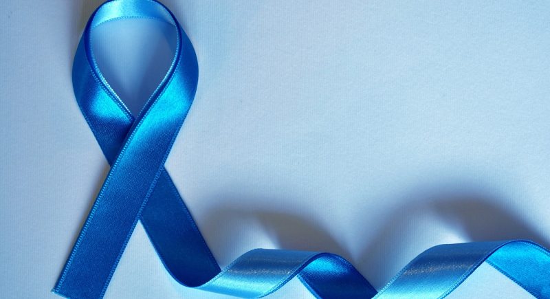 Blaues Schleifenband, zu einer symbolischen Schleife geformt, liegt auf einem hellblauen Hintergrund. Die Schleife steht für Bewusstsein und Unterstützung bestimmter Gesundheitskampagnen, wie beispielsweise Prostatakrebs- oder Darmkrebsvorsorge. Der restliche Teil des Bandes schlängelt sich sanft über die Oberfläche.