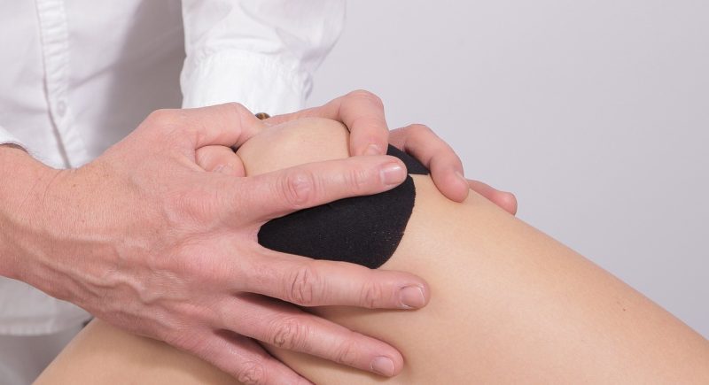 Nahaufnahme einer physiotherapeutischen Behandlung am Knie. Eine Person in einem weißen Kittel hält mit beiden Händen das Knie einer anderen Person, die eine schwarze Bandage oder ein Kinesiotape trägt. Die Szene deutet auf eine Untersuchung oder Therapie zur Schmerzlinderung oder Rehabilitation hin.