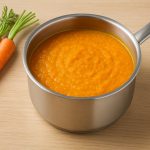 Foto einer warmen, orangefarbenen Karottensuppe in einem Edelstahltopf auf einem hellen Holzbrett. Daneben liegen drei frische Karotten mit grünem Blattansatz. Die Darstellung ist im Querformat (16:9) und zeigt eine schlichte, natürliche Szene mit Fokus auf die Suppe.