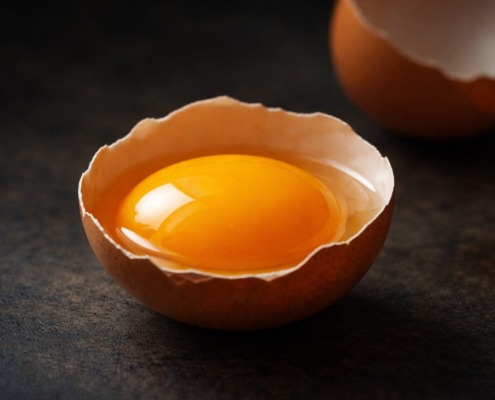 Nahaufnahme eines unversehrten Eigelbs in aufgeschlagener Eierschale vor dunklem Hintergrund, Symbol für cholinreiche Ernährung